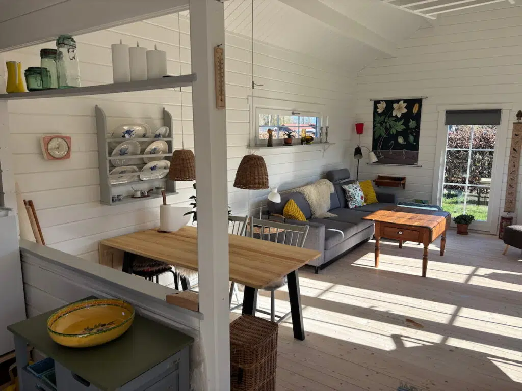 Scandinavian summerhouse interior showing lounge area with wicker pendant lights, linen cushions, and tongue and groove walls