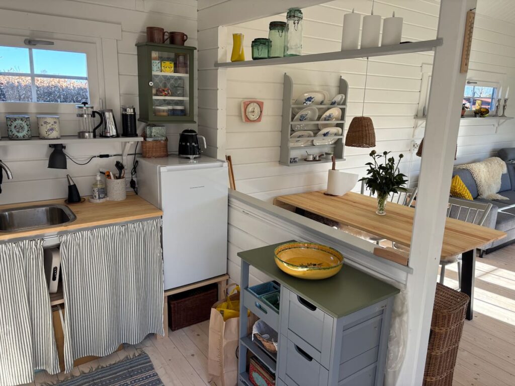 Small summerhouse kitchen with sink, under-counter fridge, and open shelving displaying ceramics and kitchenware