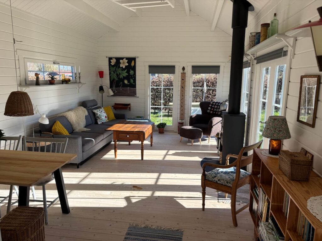 White-painted interior of a Scandinavian summerhouse with wood-burning stove, grey sofa, and exposed apex roof